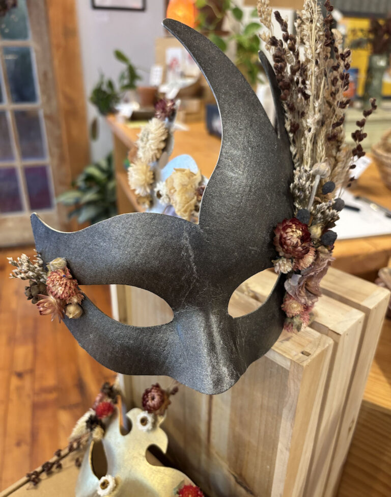 The Masquerade Mask Kit, featuring metallic details with dried flowers and grasses, rests on a wooden box in a warmly lit room with wooden floors and plants in the background.