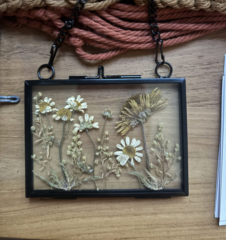 The Pressed Flower Frame features yellow and white wildflowers with greenery, set in a glass frame with a black border and chain. It is displayed on a wooden surface beside woven coral-colored yarn.
