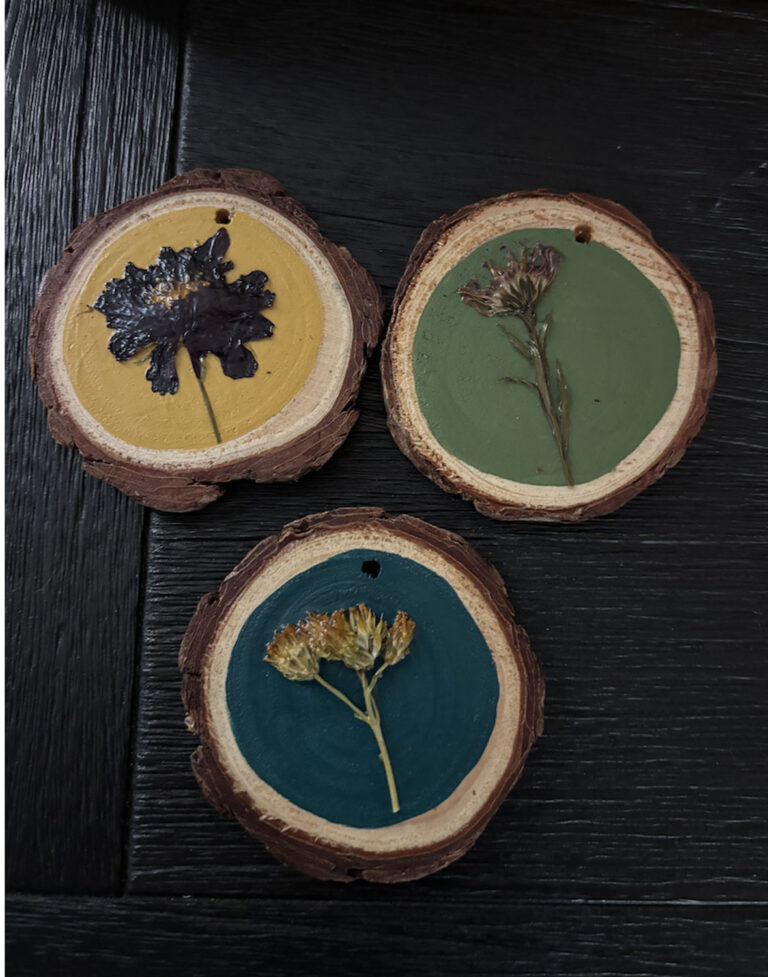 Three Pressed Flower Ornaments—round wooden slices in yellow, green, and teal—each feature a unique pressed flower or plant sealed on top and are arranged on a dark wooden surface.