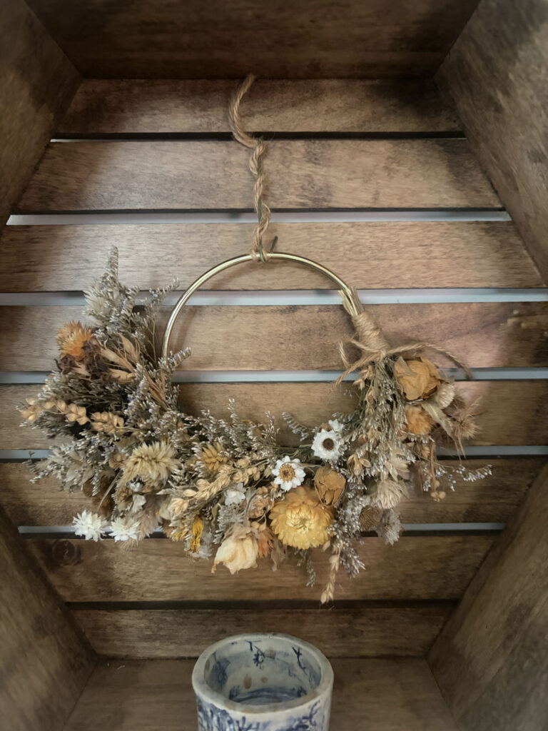 A 3"-5" Dried Floral Ring with yellow, cream, and brown flowers hangs on a wooden slatted wall. Beneath the rustic wreath, there is a small blue and white ceramic cup.