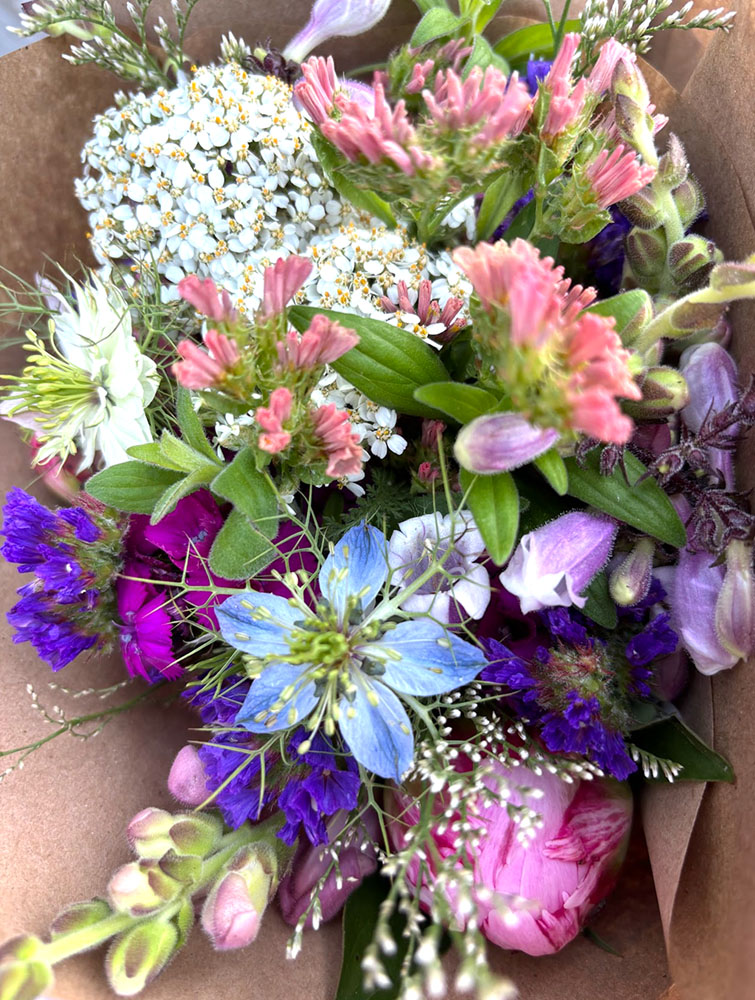 A close-up of fresh, vibrant pink, blue, purple, and white wildflowers with green foliage from the 2026 6-Week Spring Flower Subscription, delicately arranged and wrapped in brown paper for a natural look.