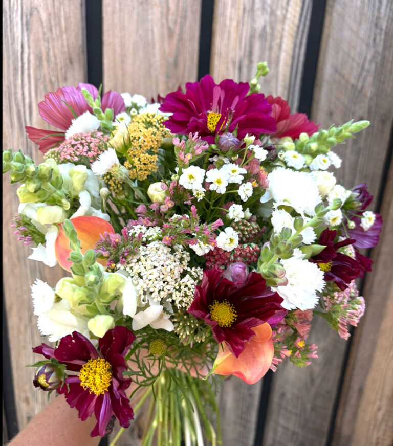 A hand holds a vibrant bouquet featured in the 2026 4-Week Summer Flower Subscription, displaying white, pink, orange, and deep magenta blooms against a rustic wooden background.