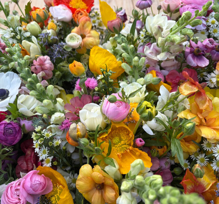 A vibrant mix of fresh, colorful flowers including yellow, pink, white, orange, and purple blooms with green stems and leaves, arranged closely together in a lush, natural bouquet.