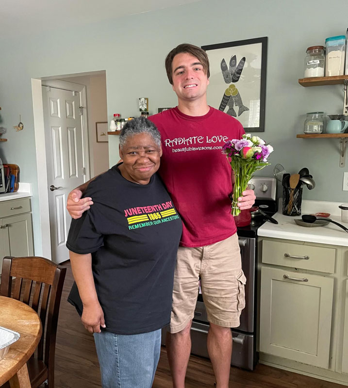 A smiling woman and man stand together in a kitchen. The man holds a bouquet of flowers and has his arm around the woman. The woman wears a Juneteenth t-shirt; the man wears a Radiate Love shirt.